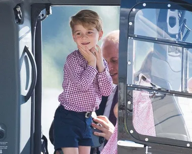 A young boy in a checkered shirt stands in a helicopter doorway, smiling with hands on his cheeks.