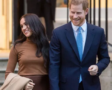 Prince Harry and Meghan Markle smiling and holding hands outside a building, both dressed in formal attire.