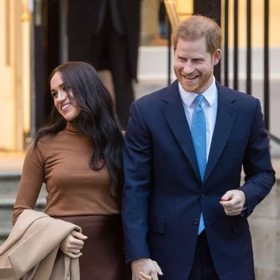 Prince Harry and Meghan Markle smiling and holding hands outside a building, both dressed in formal attire.