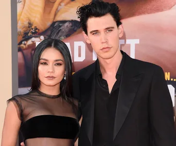 Two individuals posing at a formal event, one wearing a sheer dress and the other in a black suit, standing close together.