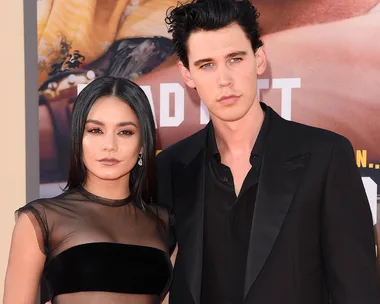 Two individuals posing at a formal event, one wearing a sheer dress and the other in a black suit, standing close together.