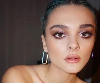 Woman with brown eyes and shimmering eyeshadow, wearing rectangular earrings and glossy nude lipstick.