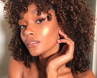 Person with curly hair showing glowing skin, highlighting cheeks, and glossy lips, posing against a neutral background.