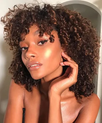 Person with curly hair showing glowing skin, highlighting cheeks, and glossy lips, posing against a neutral background.