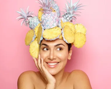 Smiling woman with a playful pineapple and sliced fruit hat against a pink background.