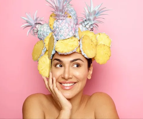 Smiling woman with a playful pineapple and sliced fruit hat against a pink background.