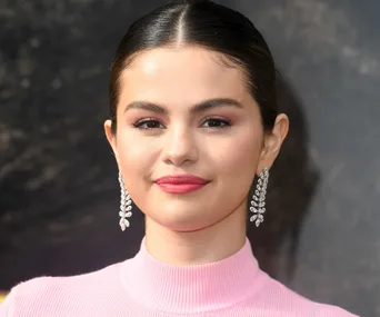 Selena Gomez at an event, wearing a pink top and diamond earrings, smiling with a neutral background.