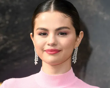 Selena Gomez at an event, wearing a pink top and diamond earrings, smiling with a neutral background.