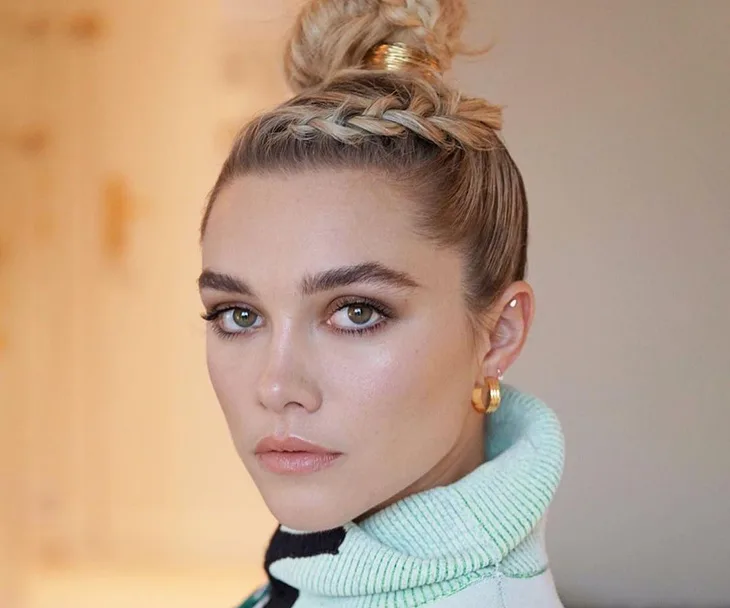 A person with braided updo hairstyle, wearing a light-colored sweater and gold hoop earrings, looking towards the camera.