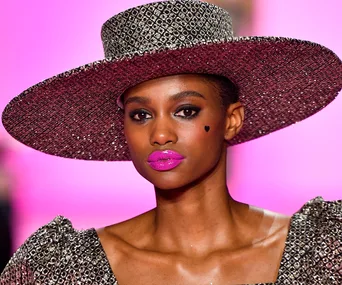 Woman in textured wide-brimmed hat, bold pink lipstick, heart face detail against a pink background. Fashion runway look.