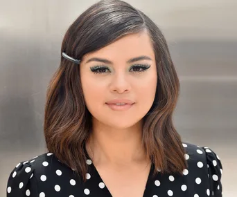 Woman with shoulder-length brown hair, wearing a black polka dot dress and hairpin, looks forward with a neutral expression.