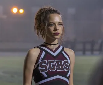 A cheerleader in an SGHS uniform stands on a field, looking serious with her hair in a ponytail and wearing dark makeup.