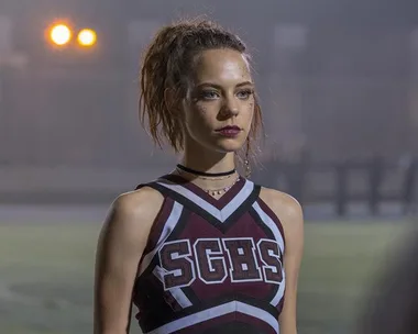 A cheerleader in an SGHS uniform stands on a field, looking serious with her hair in a ponytail and wearing dark makeup.