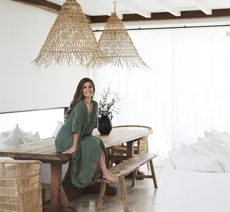 Woman in green dress sitting on a wooden table in a bright room with wicker lighting and rustic decor.