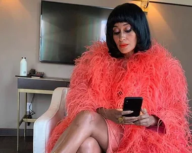 Person in a vibrant pink feathered outfit sits on a chair, looking at a smartphone.