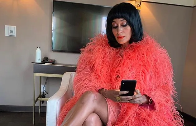 Person in a vibrant pink feathered outfit sits on a chair, looking at a smartphone.