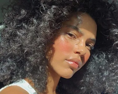 Portrait of a person with curly hair and sunlight on their face, wearing a white top.