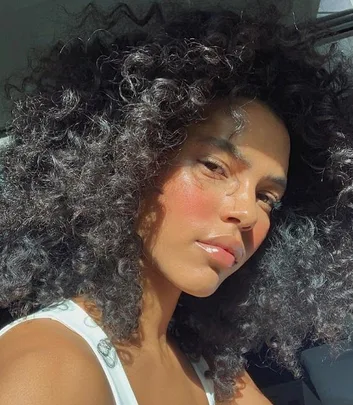 Portrait of a person with curly hair and sunlight on their face, wearing a white top.