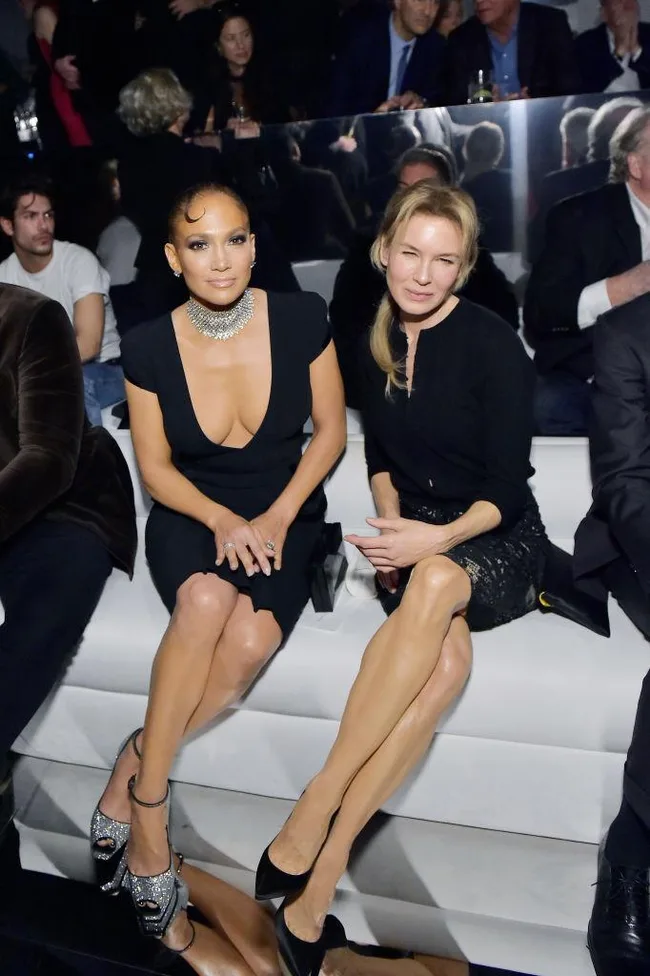 Two women sitting on a white couch at a formal event, dressed elegantly, with multiple people in the background.