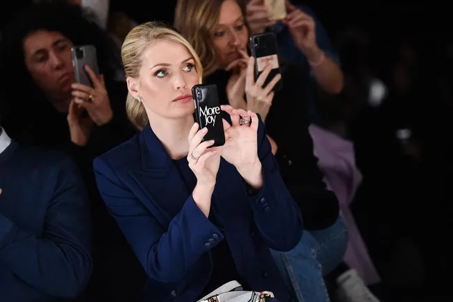 A woman in a blue blazer takes a photo with a phone case that reads "More Joy" at a fashion event.