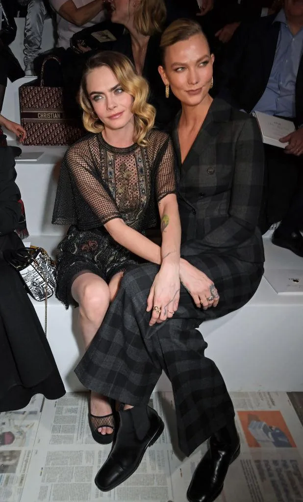 Two women sitting closely in stylish outfits at a fashion event, surrounded by other attendees.