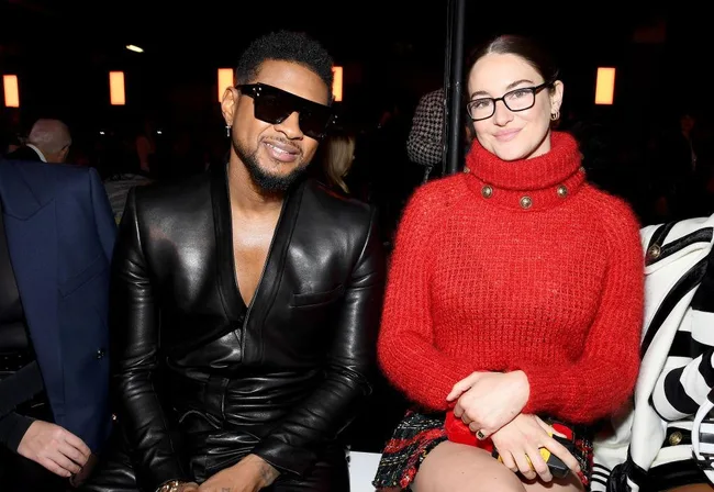 Two people seated at a fashion event, one in a black leather outfit and sunglasses, the other in a red sweater with glasses.
