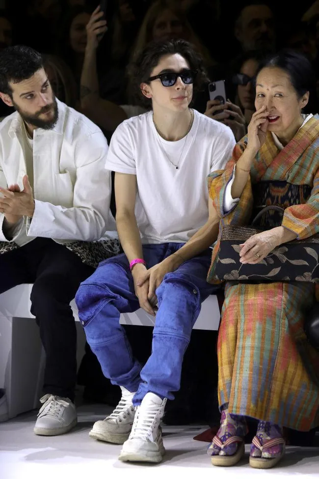 Three people sitting at a fashion show; one in sunglasses, another in a kimono, and a third person clapping.