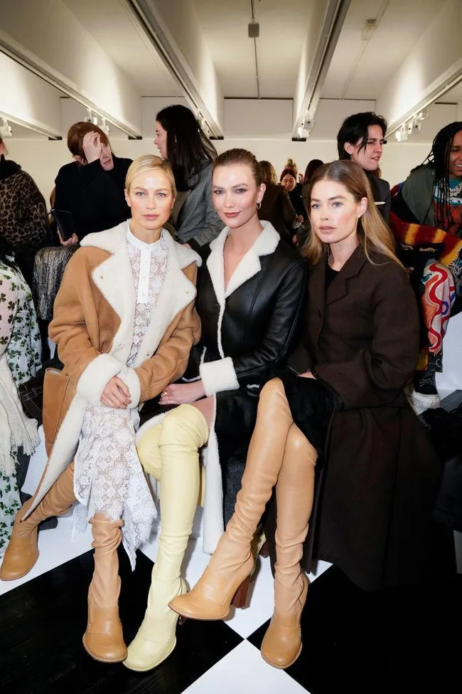 Three women sit together in fashionable coats and thigh-high boots at a runway show, surrounded by other attendees.