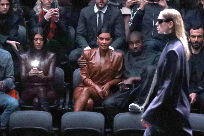 People sitting in the front row at a fashion show, with a model walking past in the foreground.