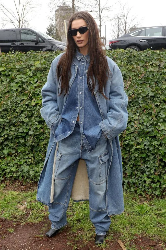 A person in oversized denim outfit and sunglasses stands outdoors with cars and greenery in the background.