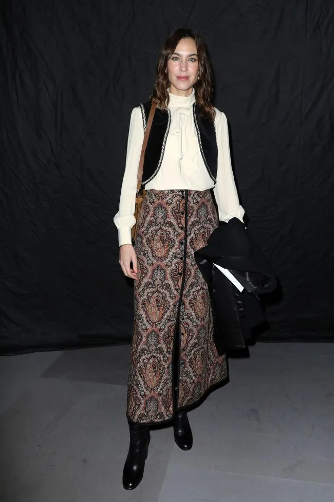 A person in a stylish outfit with a patterned skirt, cream blouse, and black boots stands against a black backdrop.