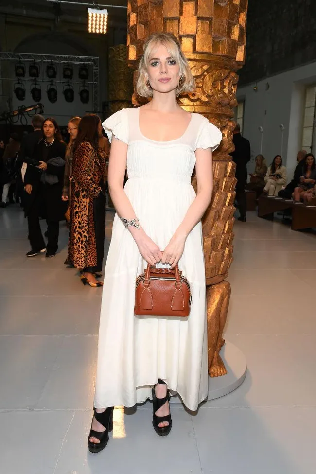 A woman in a white dress holding a brown handbag stands in front of a golden pillar at a fashion event.