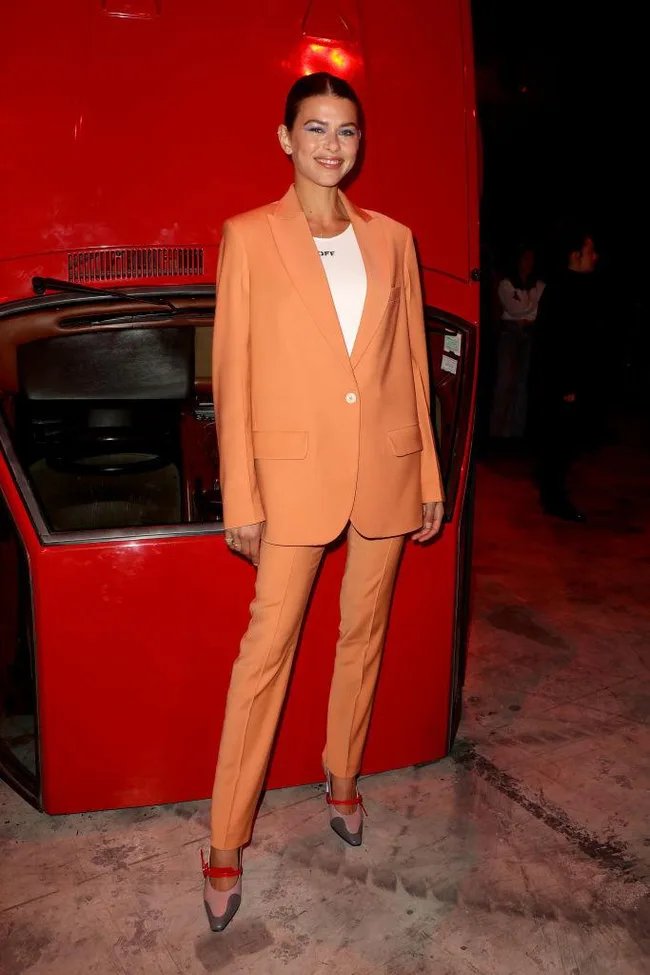 A person in an orange suit stands smiling in front of part of a red vehicle.