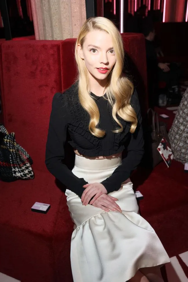 A woman with long blonde hair in a black blouse and white skirt sits on a red velvet couch, hands clasped.