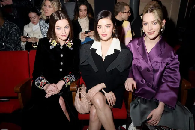 Three women seated in a theater, dressed in fashion-forward coats, surrounded by an audience.