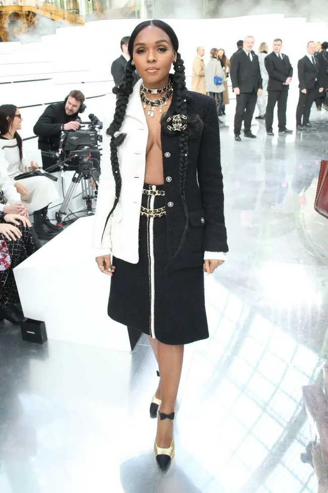 A person in a stylish black and white outfit with braided hair stands confidently on a shiny floor at a fashion event.