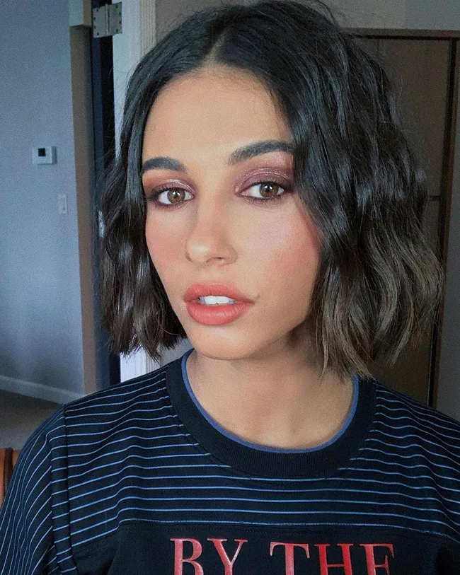 A woman with wavy dark hair and subtle makeup poses indoors, wearing a navy striped shirt.