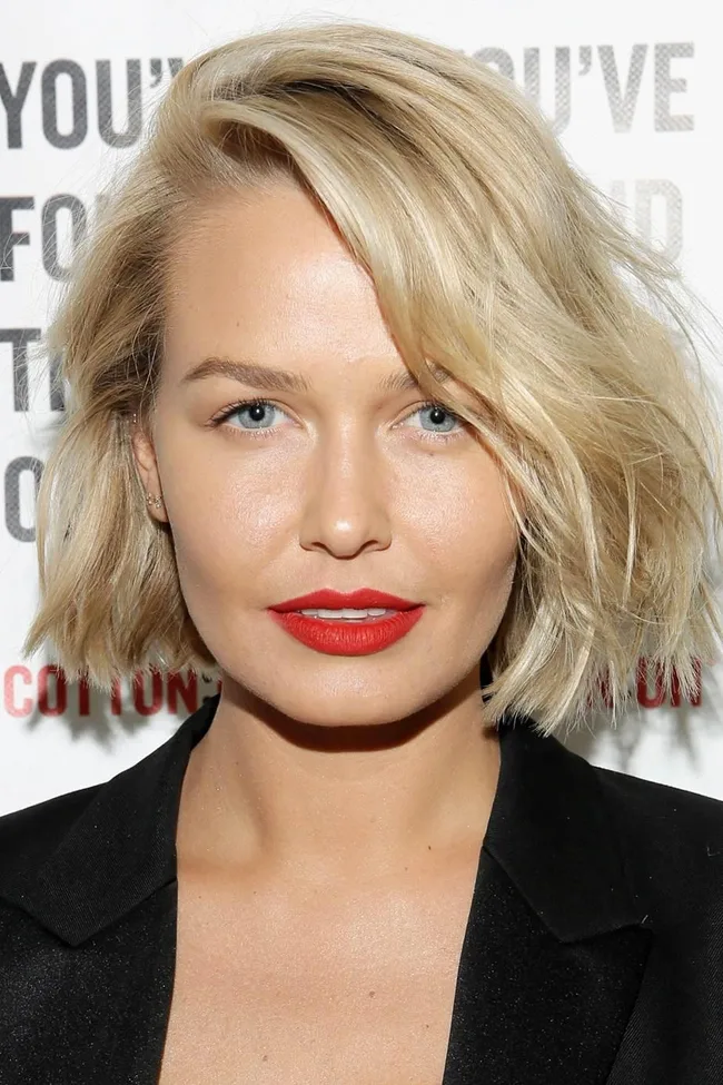 Blonde woman with red lipstick and a black blazer, standing in front of a promotional poster with text in the background.