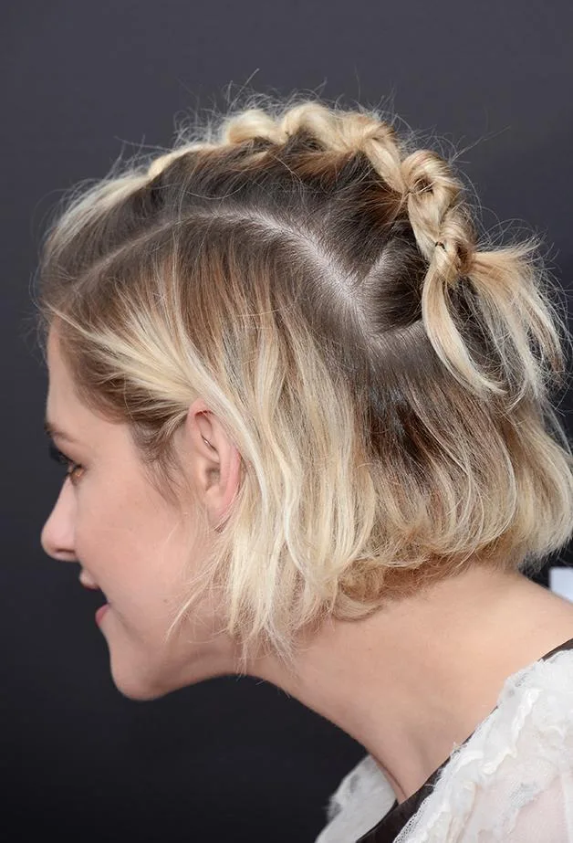 Woman with short blonde hair styled in a braided updo, side view against a dark background.