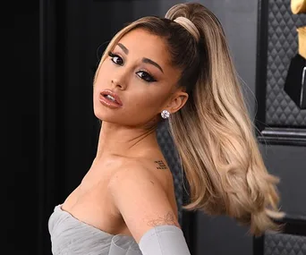 A woman with high ponytail, wearing off-shoulder dress, looking over shoulder at an event with black and gold background.