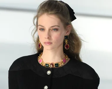 Model with long hair, wearing black quilted outfit, bold gold jewelry, on Chanel AW20 runway.