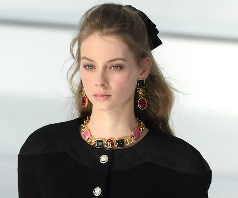 Model with long hair, wearing black quilted outfit, bold gold jewelry, on Chanel AW20 runway.