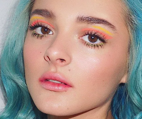 Young person with blue hair, vibrant eye makeup with orange and yellow accents, and natural lip color.