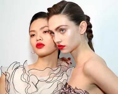 Two models with glowing skin and bold red lipstick, wearing ruffled tops, pose against a white background.