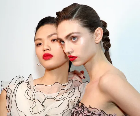 Two models with glowing skin and bold red lipstick, wearing ruffled tops, pose against a white background.