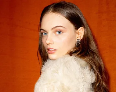 Woman with long hair wearing fur coat, posing against an orange background.