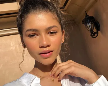 Young woman with glowing skin and curly hair in an outdoor setting, wearing a white top and delicate necklace.