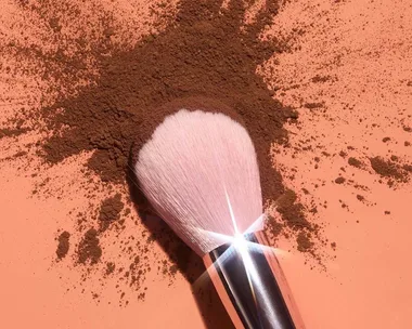 Makeup brush with white bristles and a silver handle laying on a scattered heap of brown powder on a peach surface.