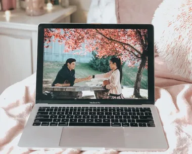 A laptop displays a scene from a movie with a man and woman exchanging an item outdoors under red-leafed trees.