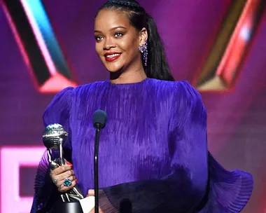 Singer in a purple dress smiles while holding a silver trophy on stage against a colorful backdrop.